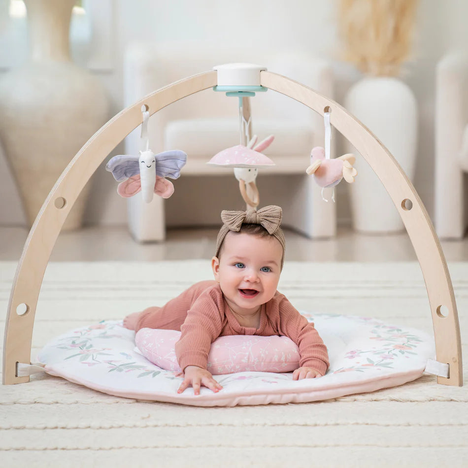Baby lying on tummy under wooden arch with hanging pastel soft toys on the Ma Fleur Activity Gym, featuring a floral patterned play mat in a cozy nursery setting.