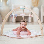 Baby lying on tummy under wooden arch with hanging pastel soft toys on the Ma Fleur Activity Gym, featuring a floral patterned play mat in a cozy nursery setting.