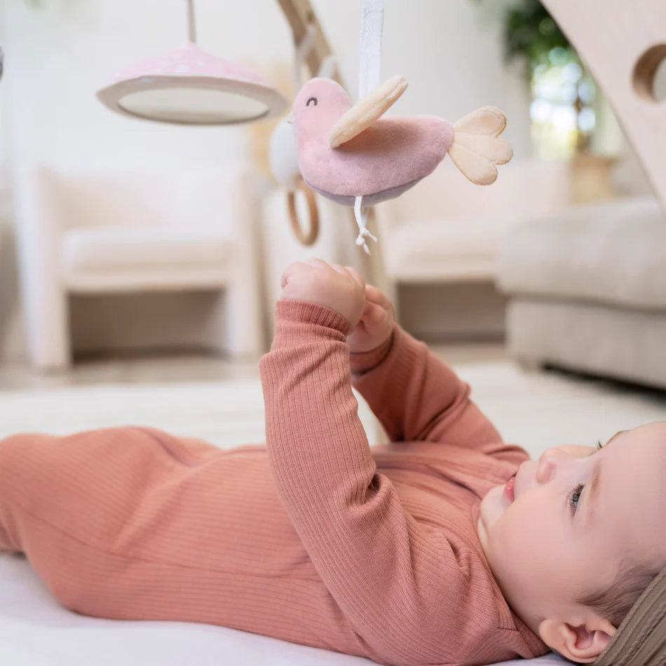 Baby lying on back reaching towards a soft pink bird toy hanging from the Ma Fleur Activity Gym in a cozy nursery setting.