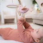 Baby lying on back reaching towards a soft pink bird toy hanging from the Ma Fleur Activity Gym in a cozy nursery setting.