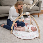 A smiling woman watches a baby lying on a floral-patterned mat under a wooden Ma Fleur Activity Gym with soft hanging toys in a cozy living room setting.