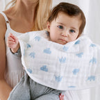 Baby wearing Animal Kingdom 2-pack Burpy Bibs featuring soft, organic muslin fabric with light blue animal prints, held by a woman in a white tank top.