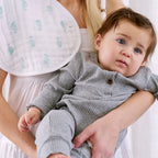 Mother holding a baby wearing a gray outfit with an Animal Kingdom 2-pack Burpy Bibs made from soft organic muslin fabric draped over her shoulder.