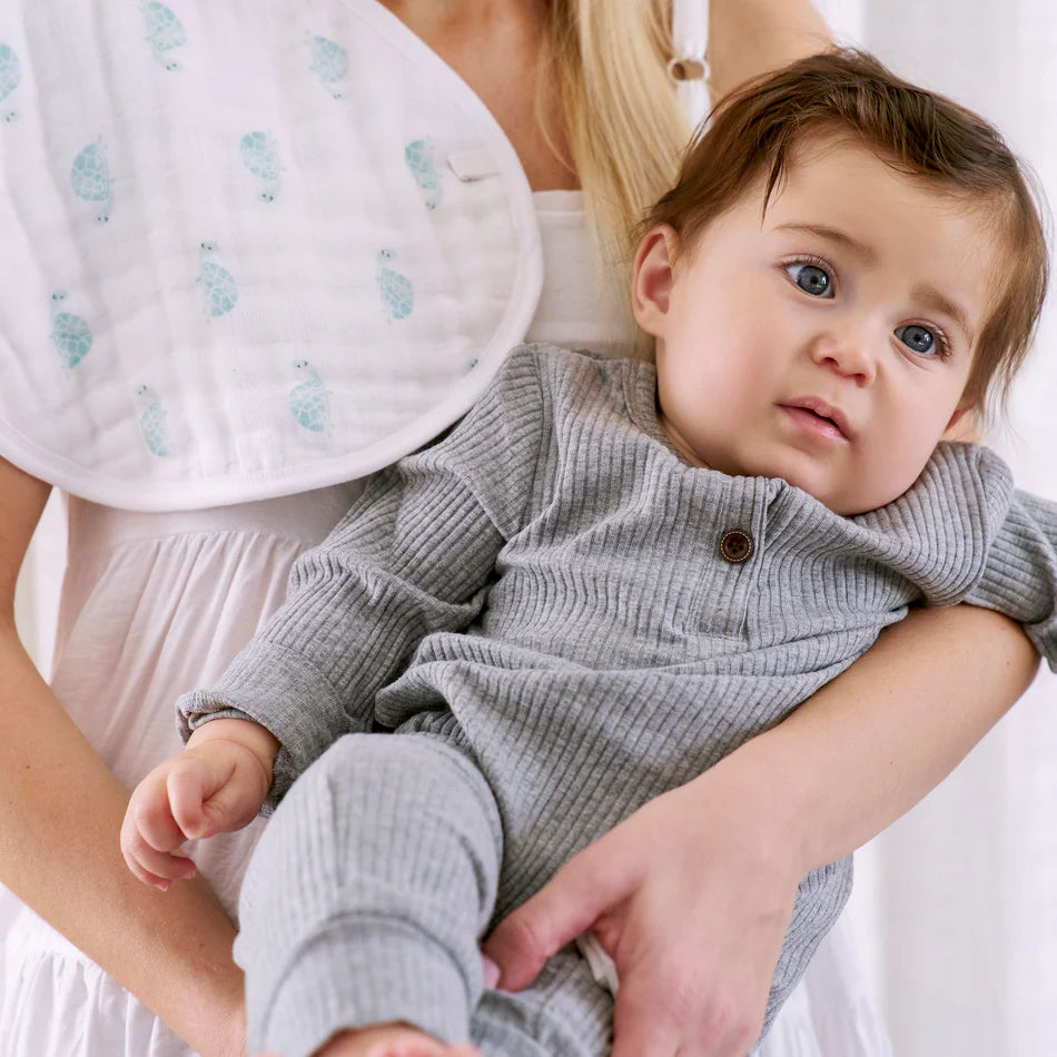 Mother holding a baby wearing a gray outfit with an Animal Kingdom 2-pack Burpy Bibs made from soft organic muslin fabric draped over her shoulder.