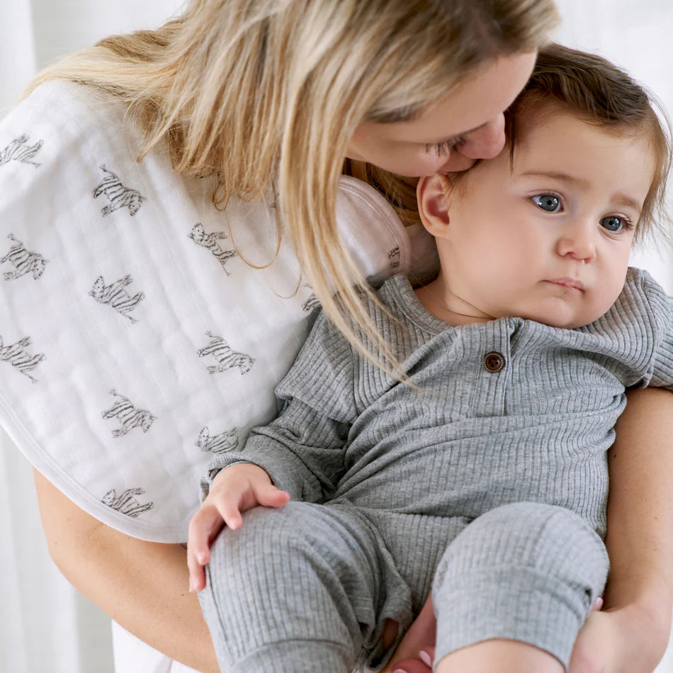 Mother holding baby wearing gray outfit and white Animal Kingdom 2-pack Burpy Bibs with zebra prints, showcasing soft, organic muslin baby gear for feeding and drooling.