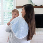 Mother holding baby over her shoulder with a light blue Rising Star 2-pack Classic Burpy Bibs featuring a subtle star pattern, in a bright living room setting.