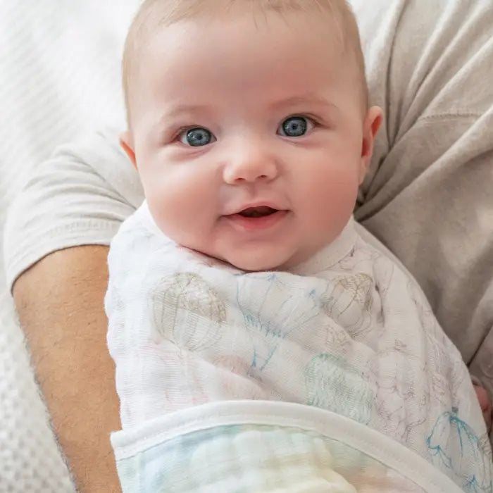 Smiling baby wrapped in soft muslin fabric wearing Above the Clouds 2-Pack Organic Burpy Bibs with a hot air balloon pattern while being held gently.