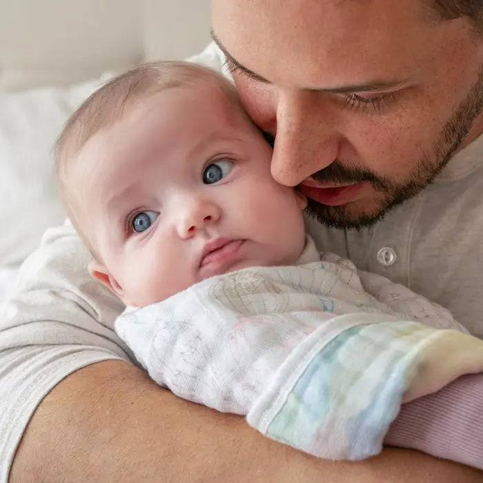 Father gently holding baby wrapped in a soft muslin cloth featuring the Above the Clouds 2-Pack Organic Burpy Bibs, showcasing comfort and care for newborn essentials.