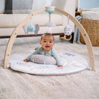 Baby lying on tummy on a soft play mat with wooden arches and hanging toys, featuring the Rising Star Play + Discovery Activity Gym in a cozy living room setting.