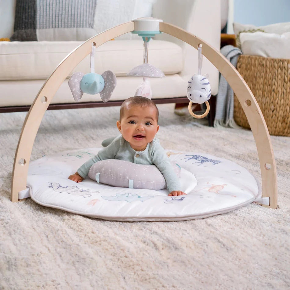 Baby lying on tummy on a soft play mat with wooden arches and hanging toys, featuring the Rising Star Play + Discovery Activity Gym in a cozy living room setting.